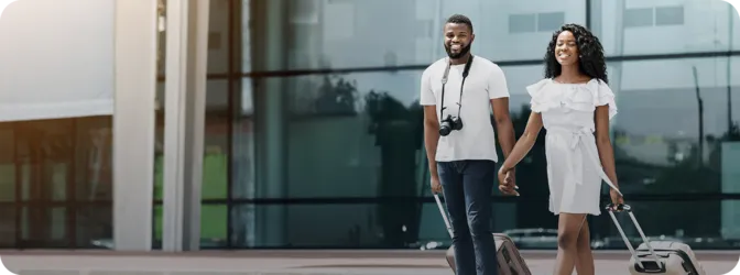 Young couple holding hands and pulling suitcases outside modern glass building.