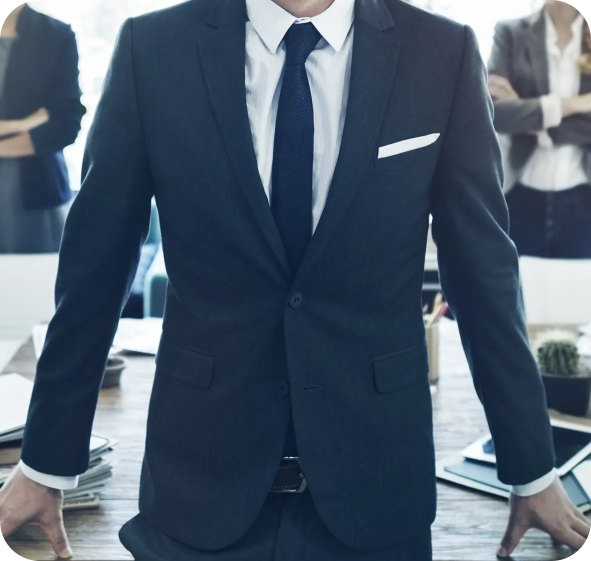 Man in dark suit and tie standing at table with two blurred businesspeople behind.