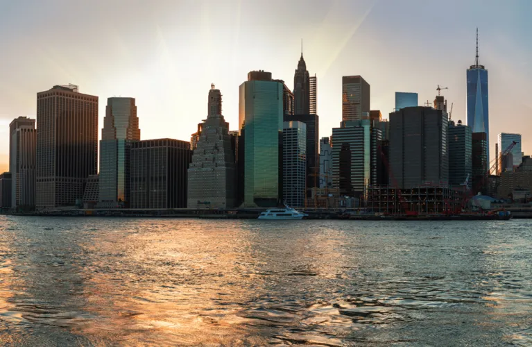 Panoramic view of New York City skyline at sunset with water foreground and illuminated skyscrapers.