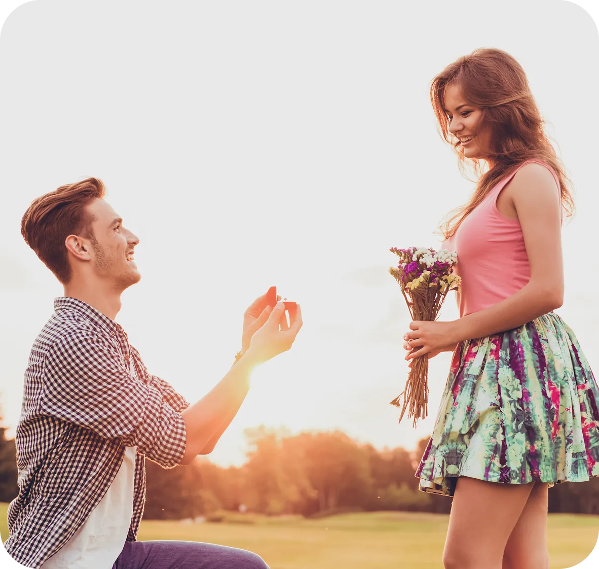Young man proposing with a ring to a smiling woman holding a bouquet outdoors at sunset.