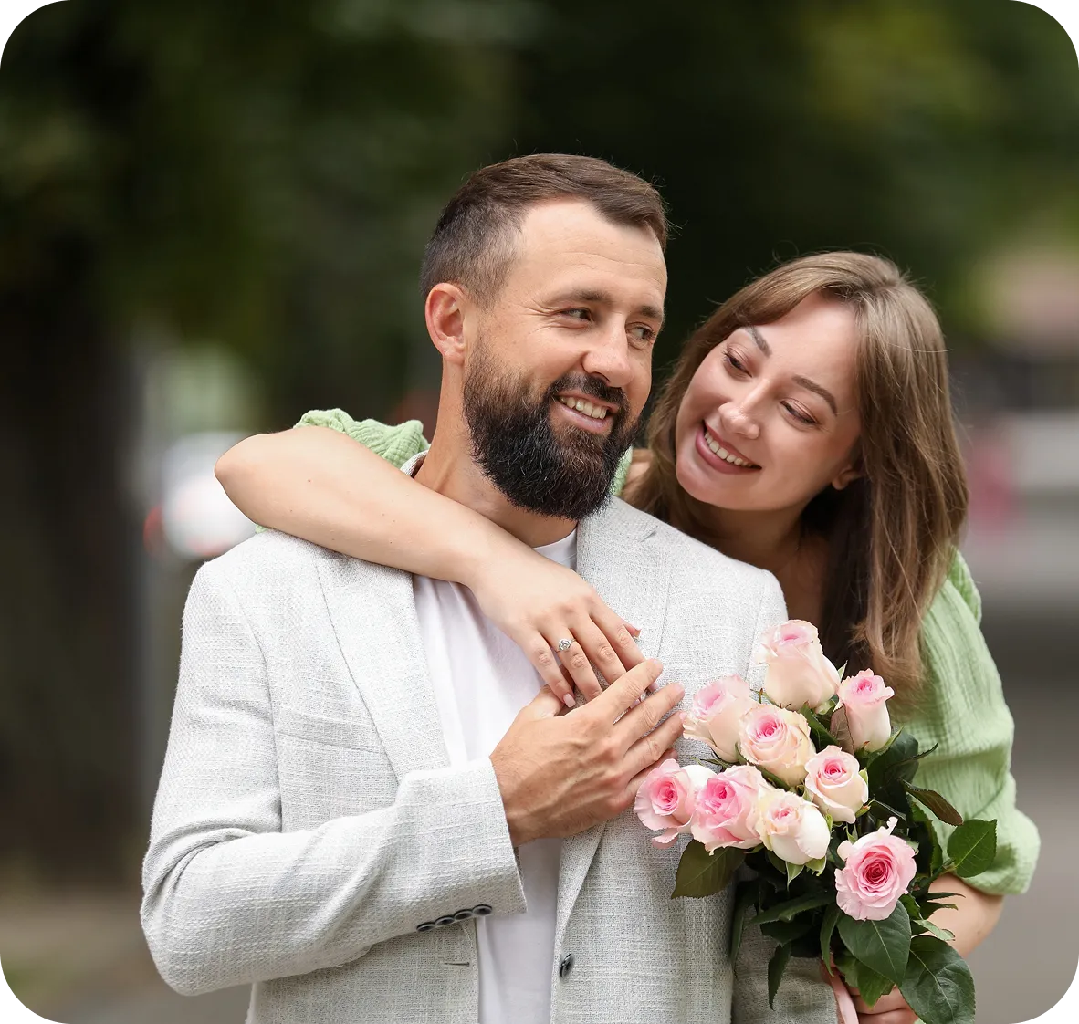 Smiling couple outdoors, woman in green dress hugging man in light gray blazer holding pink roses.