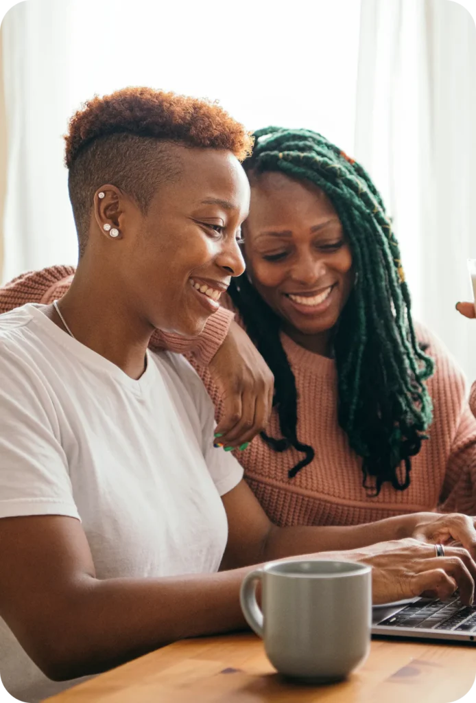 Two smiling individuals, one with short curly hair and white shirt, the other with long green braids and pink sweater, working on a laptop at a wooden table with a gray mug.