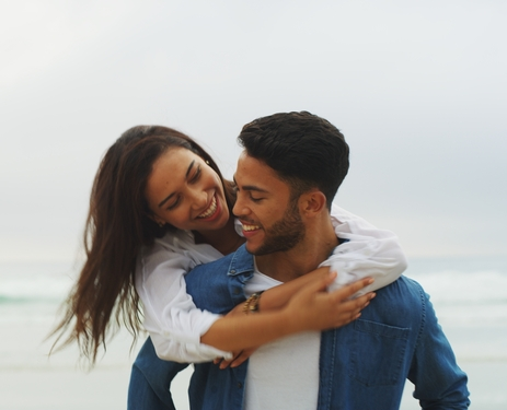 Young woman in white shirt hugging smiling man in blue jacket at beach.