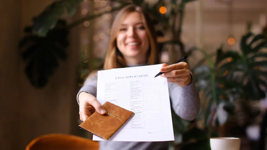 Young woman holding a visa application form and a brown passport cover, smiling indoors.