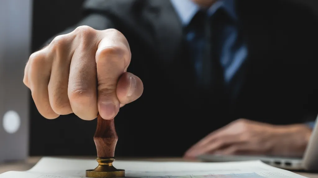 Close-up of a person stamping a document on a desk, wearing a dark suit.