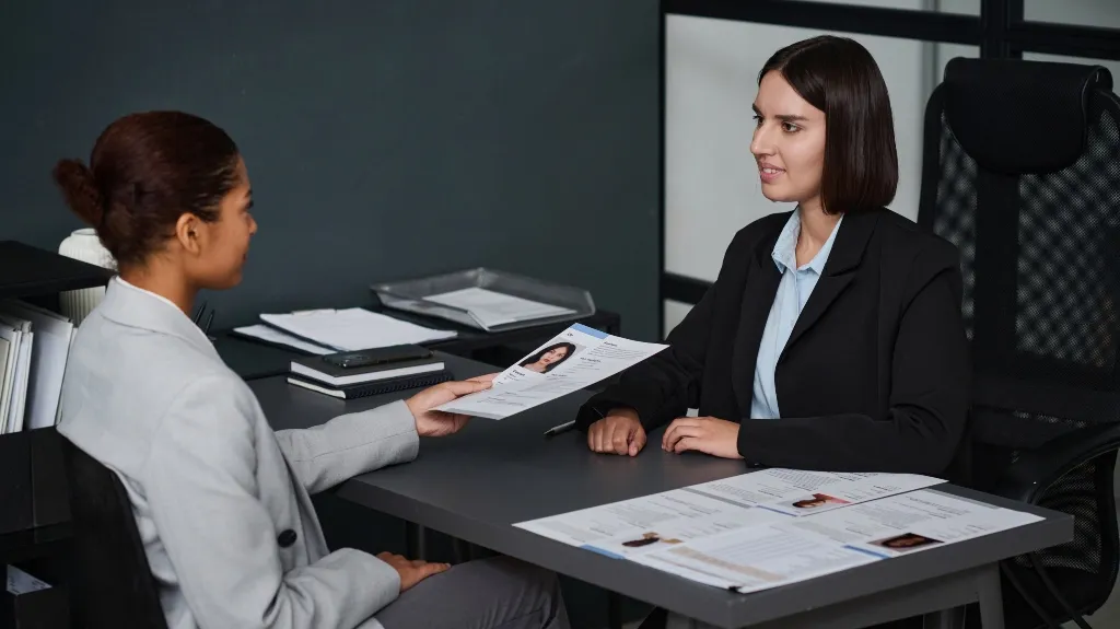 Two professional women in business attire conducting a job interview with resumes on a dark office desk.