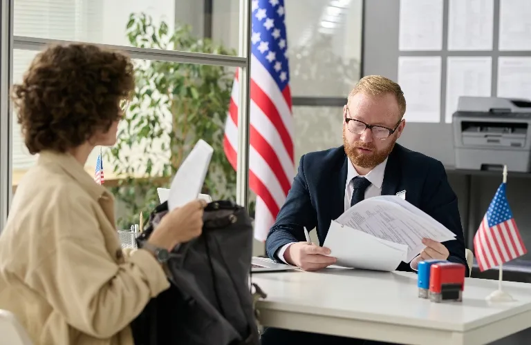 Man in suit reviewing documents at desk with American flags, woman holding papers opposite.