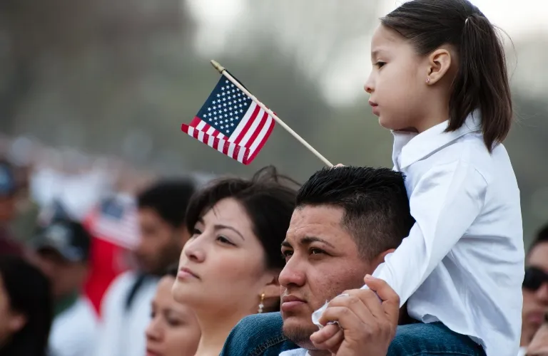 A young girl in white shirt holding an American flag seated on a man's shoulders in a crowd.