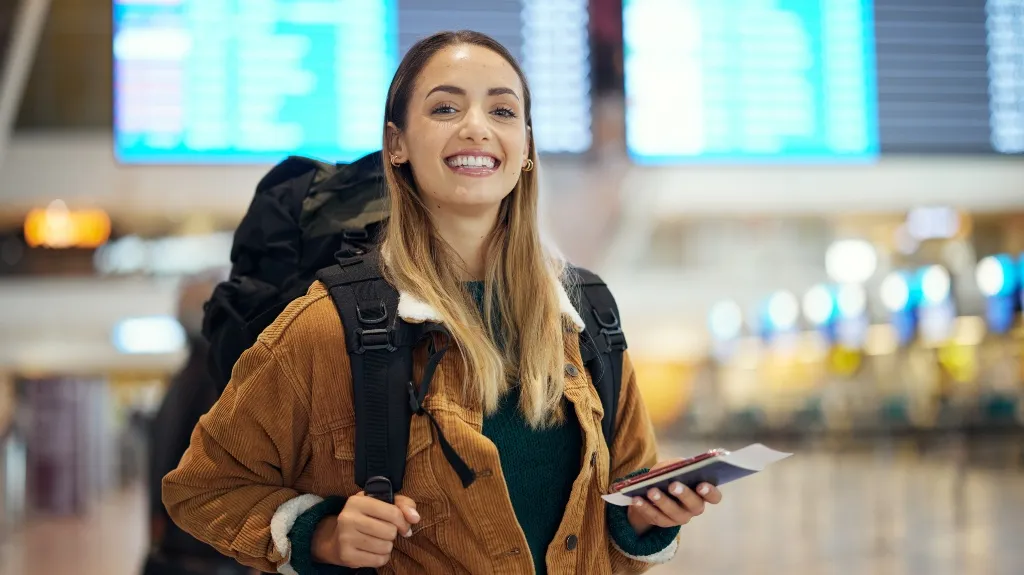 Young woman in brown jacket with backpack holding passport and boarding pass at airport.