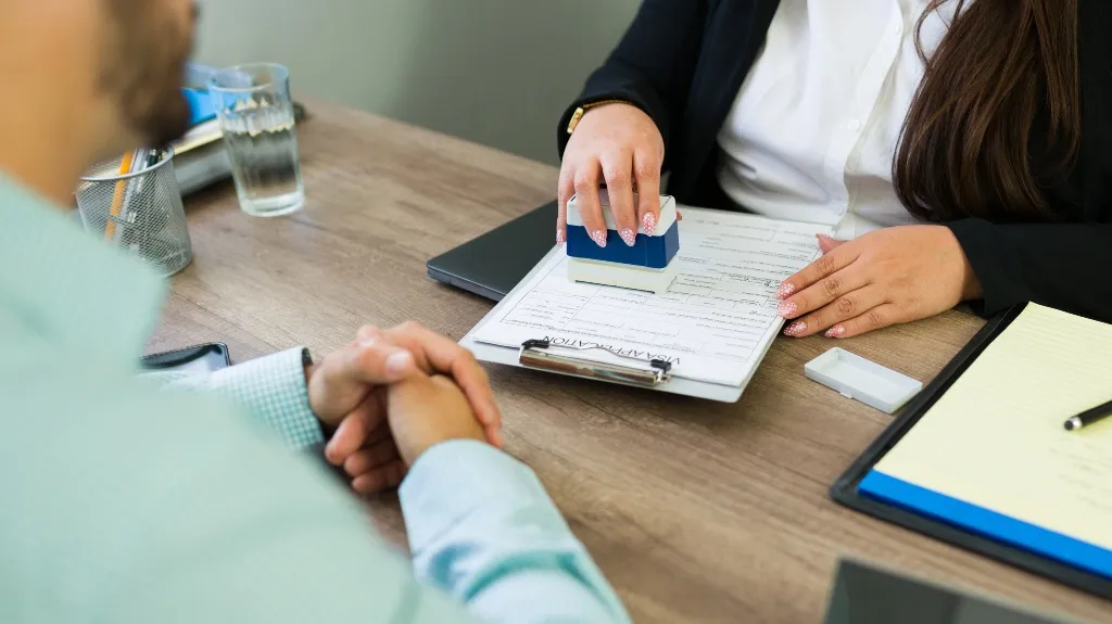 Two professionals at a desk, one stamping a visa application form on a clipboard.