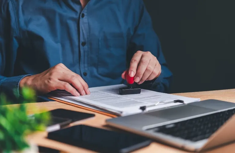 Person in blue shirt stamping a document on a desk with laptop and phone nearby.