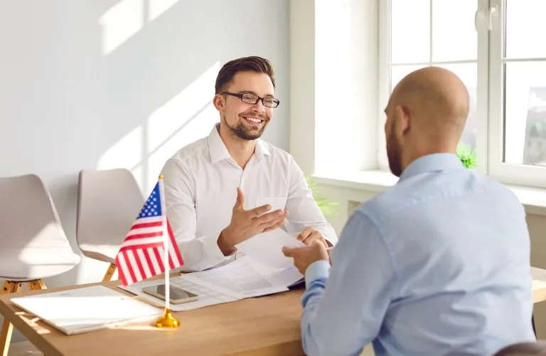Two professional men in business attire conversing across a wooden desk with an American flag on it.