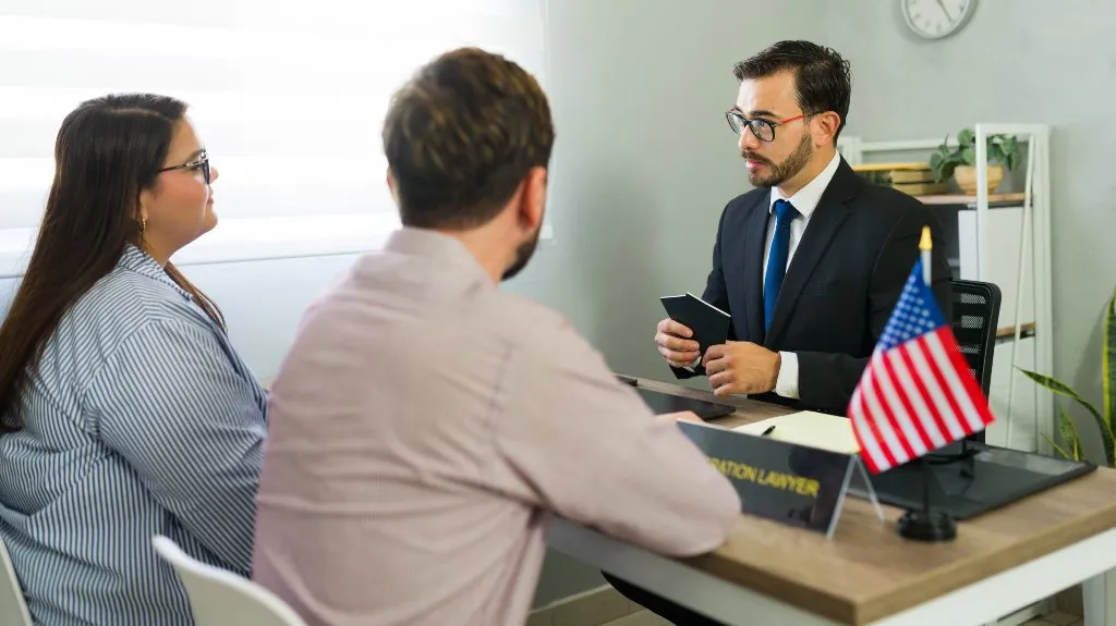 Immigration lawyer in suit consulting a couple across a desk with American flag.