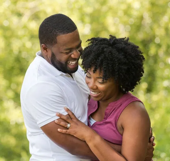 Smiling African American couple embracing outdoors with blurred green foliage background.