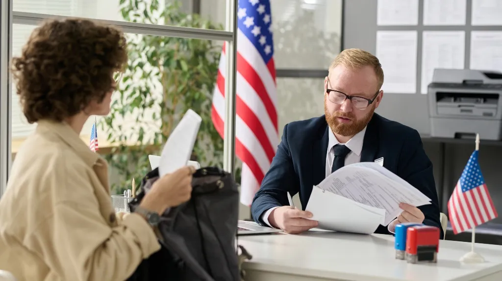 Man in suit reviewing documents across desk from seated woman in office with American flags.