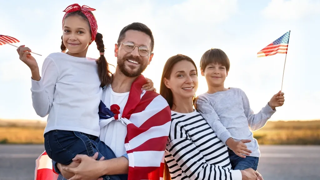 Family of four smiling outdoors, holding and wrapped in American flags at sunset.