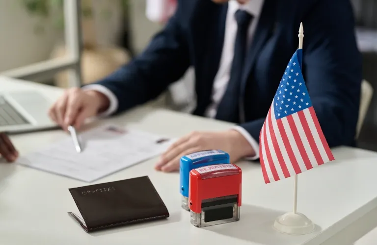 Small American flag, passport, and rejection stamps on desk with person in business attire.