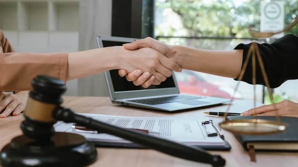Two individuals shaking hands over a desk with a gavel, legal documents, and a laptop.