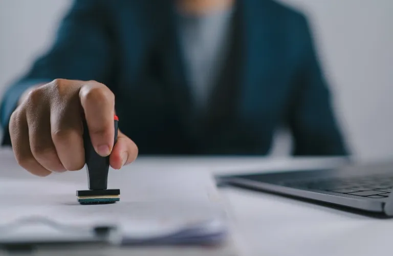 Person in dark suit stamping a document on a white desk beside a laptop.