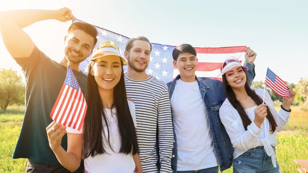 Group of five diverse young adults smiling outdoors, holding and displaying American flags.
