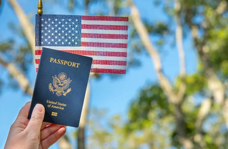 Hand holding a United States passport in front of a small American flag outdoors.