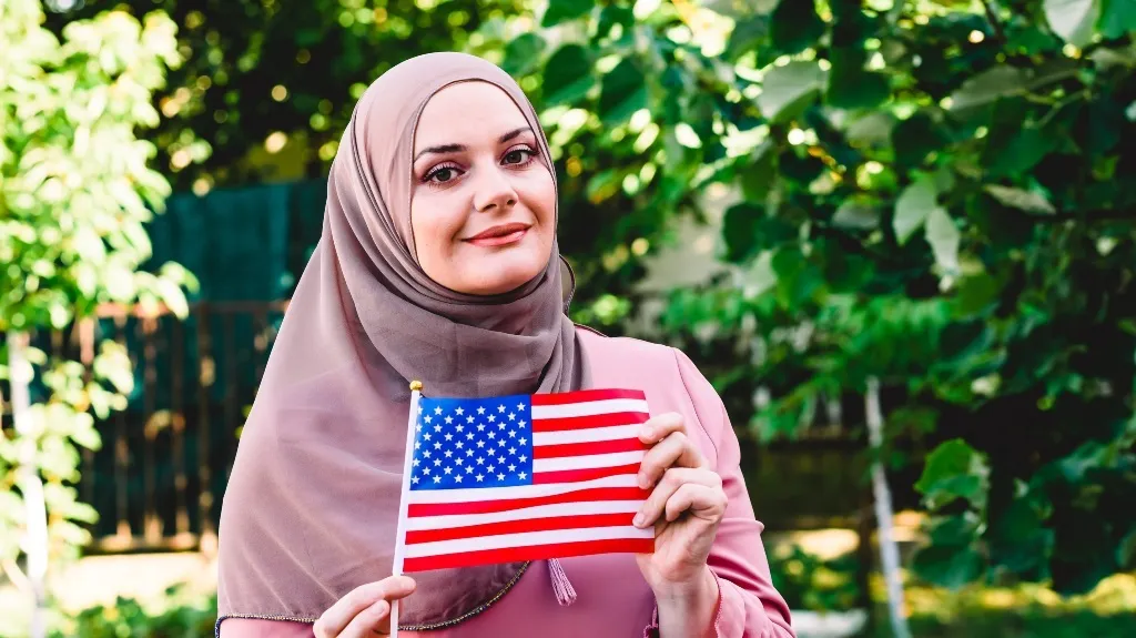 Woman wearing a mauve hijab and blouse holding a small American flag outdoors.