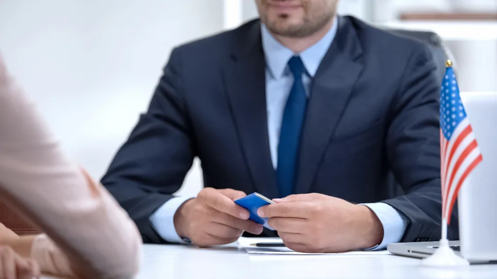 Professional man in navy suit holding a blue passport during a consultation, with a small American flag on the desk.